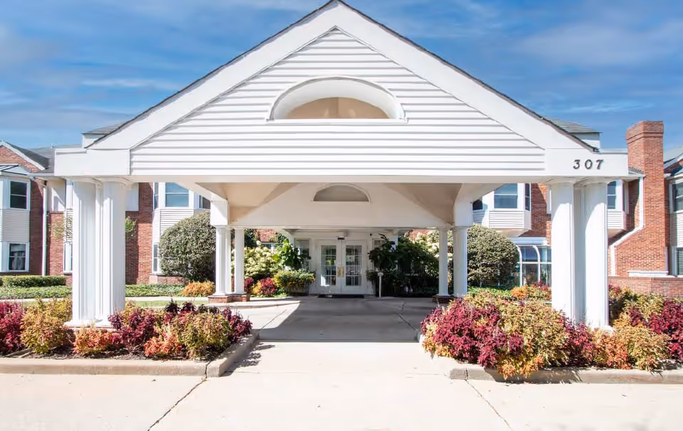 Entrance of a senior living facility with a white covered driveway supported by columns, surrounded by landscaped bushes and plants, and a brick building in the background under a blue sky.