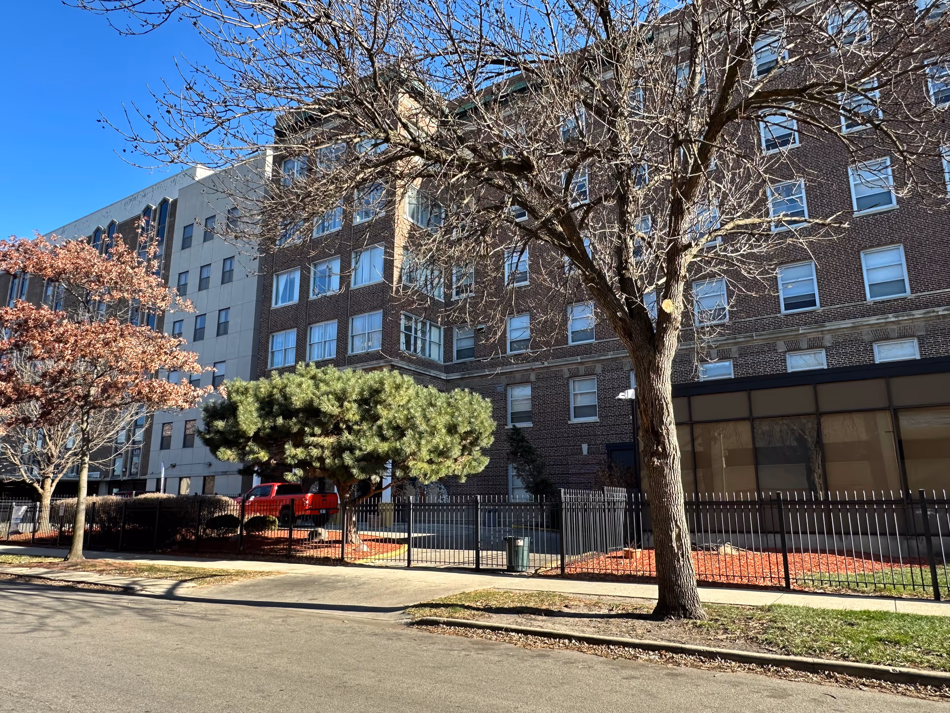 Exterior view of a multi-story brick and concrete building with many windows, surrounded by leafless and partially leafed trees, a black metal fence, and a sidewalk under a clear blue sky.