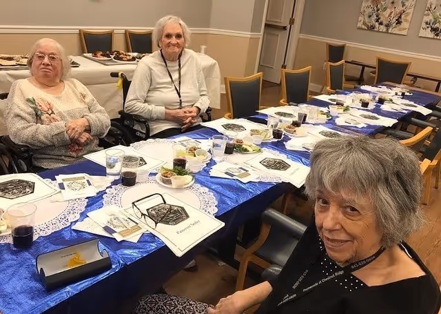 Three elderly women sitting at a long dining table covered with a blue tablecloth and white doilies, with plates of food and drinks in front of them in a well-lit room.