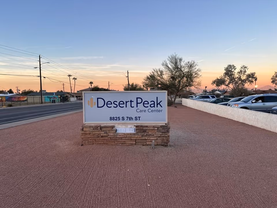 Outdoor view of a sign for Desert Peak Care Center located at 8825 S 7th Street, set on a stone base with a gravel ground and a parking lot with cars to the right. Trees and a road with power lines are visible in the background under a clear sky at sunset.