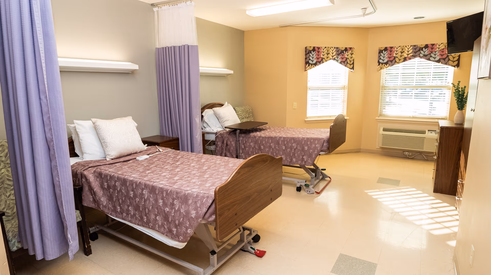 A bright and clean healthcare facility room with two hospital beds separated by a purple privacy curtain. Each bed has a patterned purple bedspread and white pillows. The room features two windows with blinds and decorative valances, a wall-mounted TV, an air conditioning unit, and a wooden dresser with a vase of flowers.