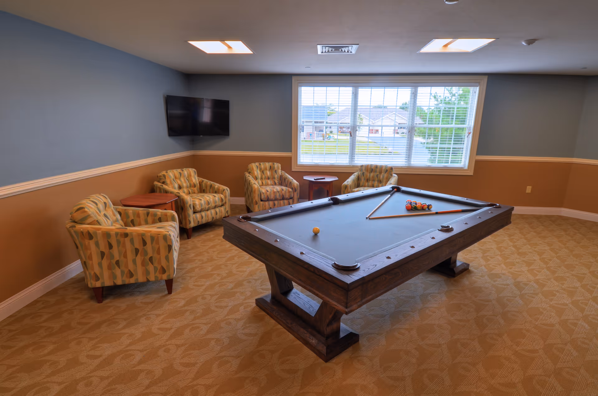 A recreational room with a pool table in the center, four patterned armchairs arranged around two small wooden side tables, a wall-mounted flat screen TV, and a large window letting in natural light. The walls are painted blue and tan with white trim, and the floor is carpeted with a patterned design.