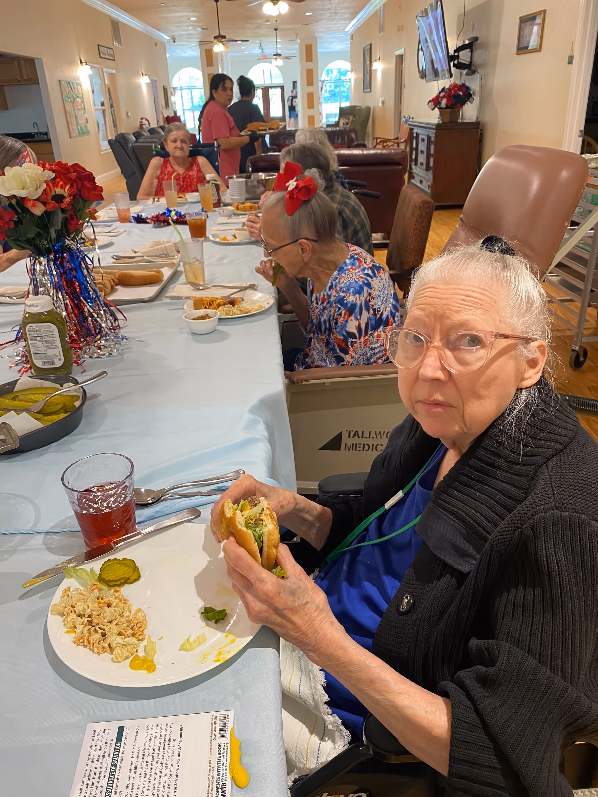 Several elderly women seated around a long dining table in a communal dining area, eating a meal that includes sandwiches, pasta salad, and pickles. One woman in the foreground is holding a sandwich and looking toward the camera. The room has a warm, inviting atmosphere with light-colored walls, ceiling fans, and a television mounted on the wall. A staff member is serving food in the background.