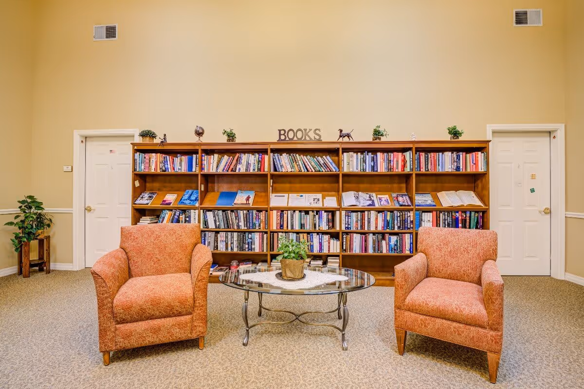 A cozy reading area in a senior living facility featuring two orange upholstered armchairs facing a glass coffee table with a potted plant. Behind the chairs is a large wooden bookshelf filled with books and decorative items, with the word 'BOOKS' displayed on top. The room has beige walls, carpeted floor, and two white doors on either side of the bookshelf.