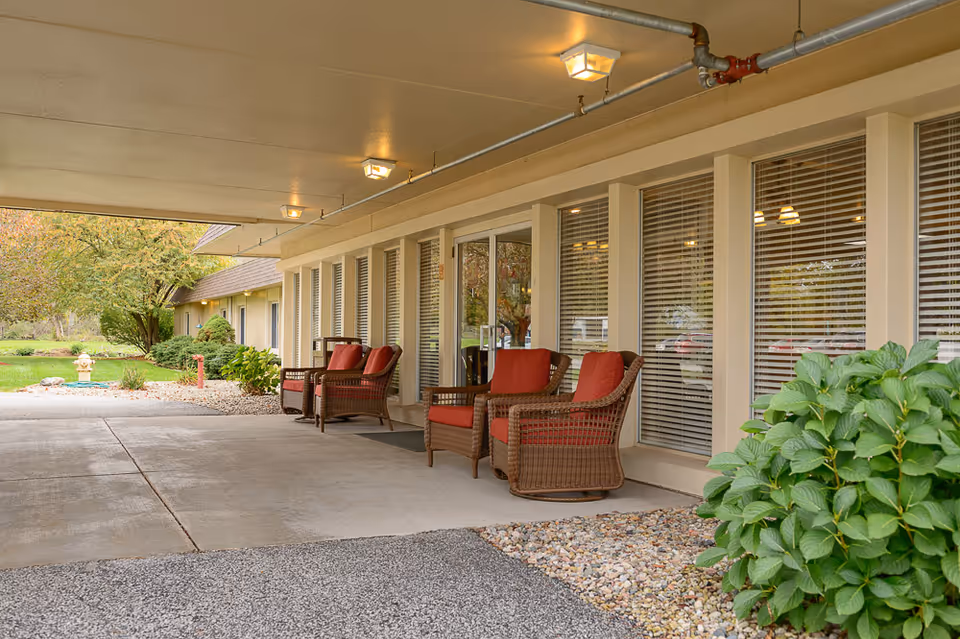 Covered entrance/portico with wicker chairs with red cushions along windows and landscaped grounds.