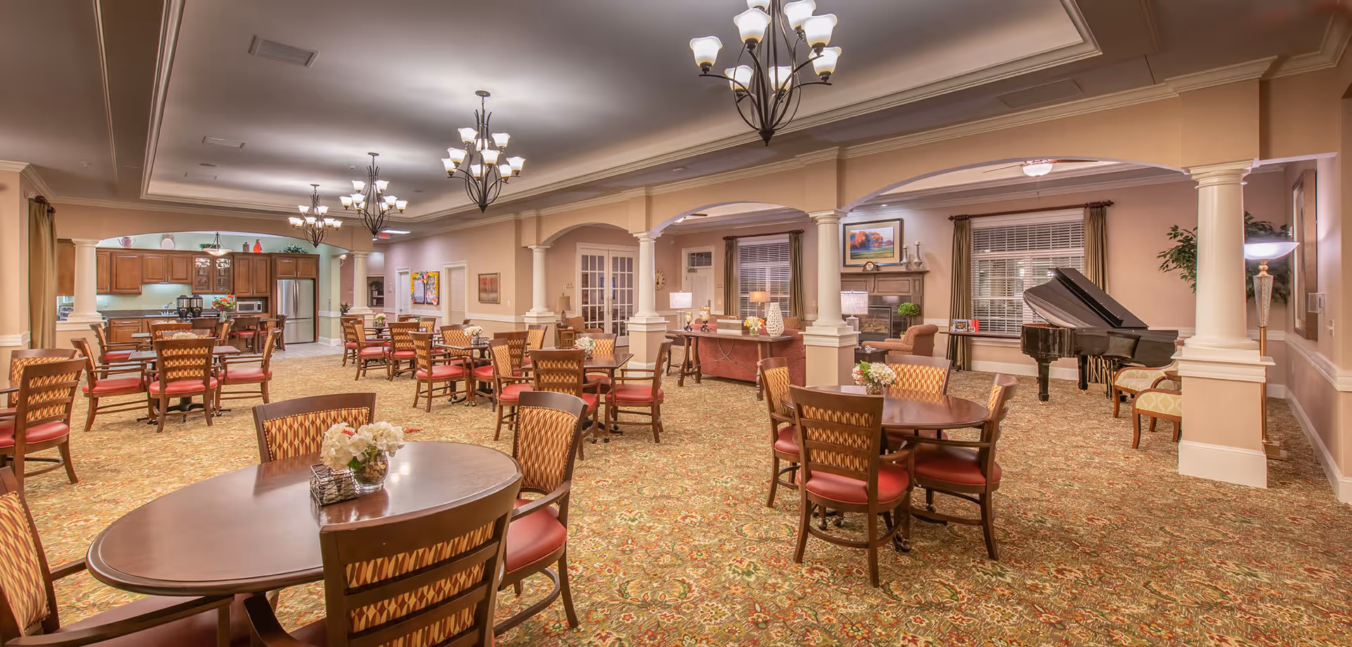 Spacious senior living dining and common area with multiple round tables and chairs, chandeliers, a grand piano, and a kitchenette in the background.