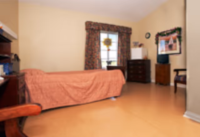 A simple bedroom with a single bed covered in a peach-colored bedspread, a wooden chair, a window with floral curtains, a dark wooden dresser, a small white refrigerator, a TV on a stand, and a framed picture on the wall.