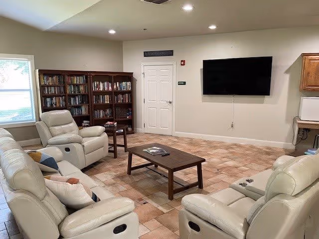 A cozy living room area with cream-colored leather recliners arranged around a wooden coffee table. There is a large flat-screen TV mounted on the wall, a bookshelf filled with books, and a window letting in natural light. The floor is tiled with a warm-toned pattern.