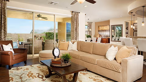 Spacious living room with a large beige sectional sofa adorned with white and beige pillows, a wooden coffee table with decorative items including a clock and a plant, a brown leather armchair, and a patterned area rug. The room has large sliding glass doors leading to a covered patio with outdoor seating and ceiling fans. In the background, there is a dining area with a table and chairs, pendant lighting, and artwork on the walls.