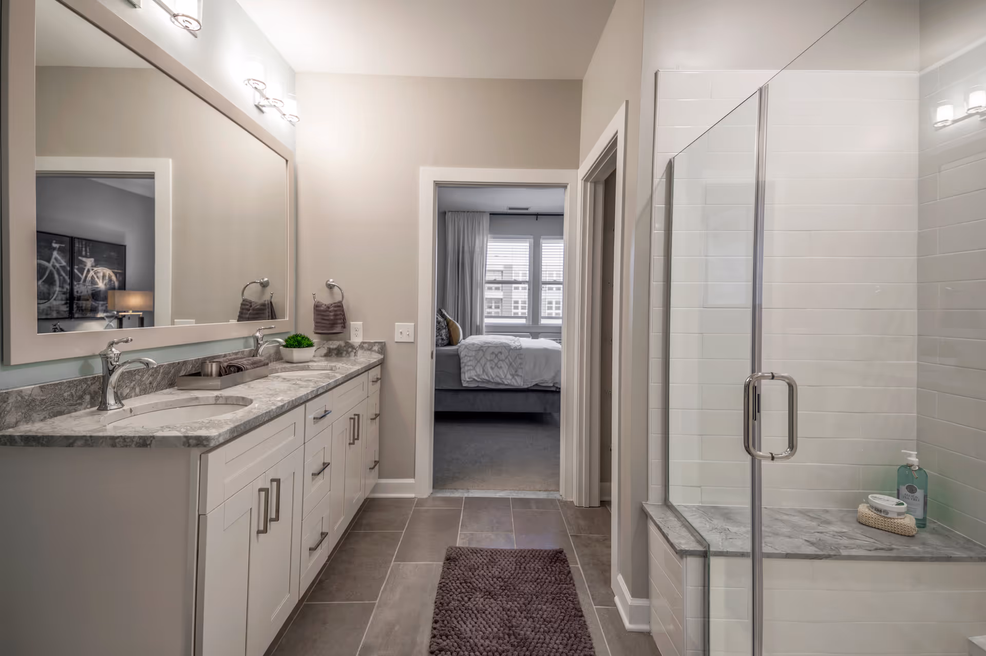 Modern bathroom with a large mirror above a double sink vanity with marble countertops. There is a glass-enclosed shower on the right and a doorway leading to a bedroom with a bed and window in the background.