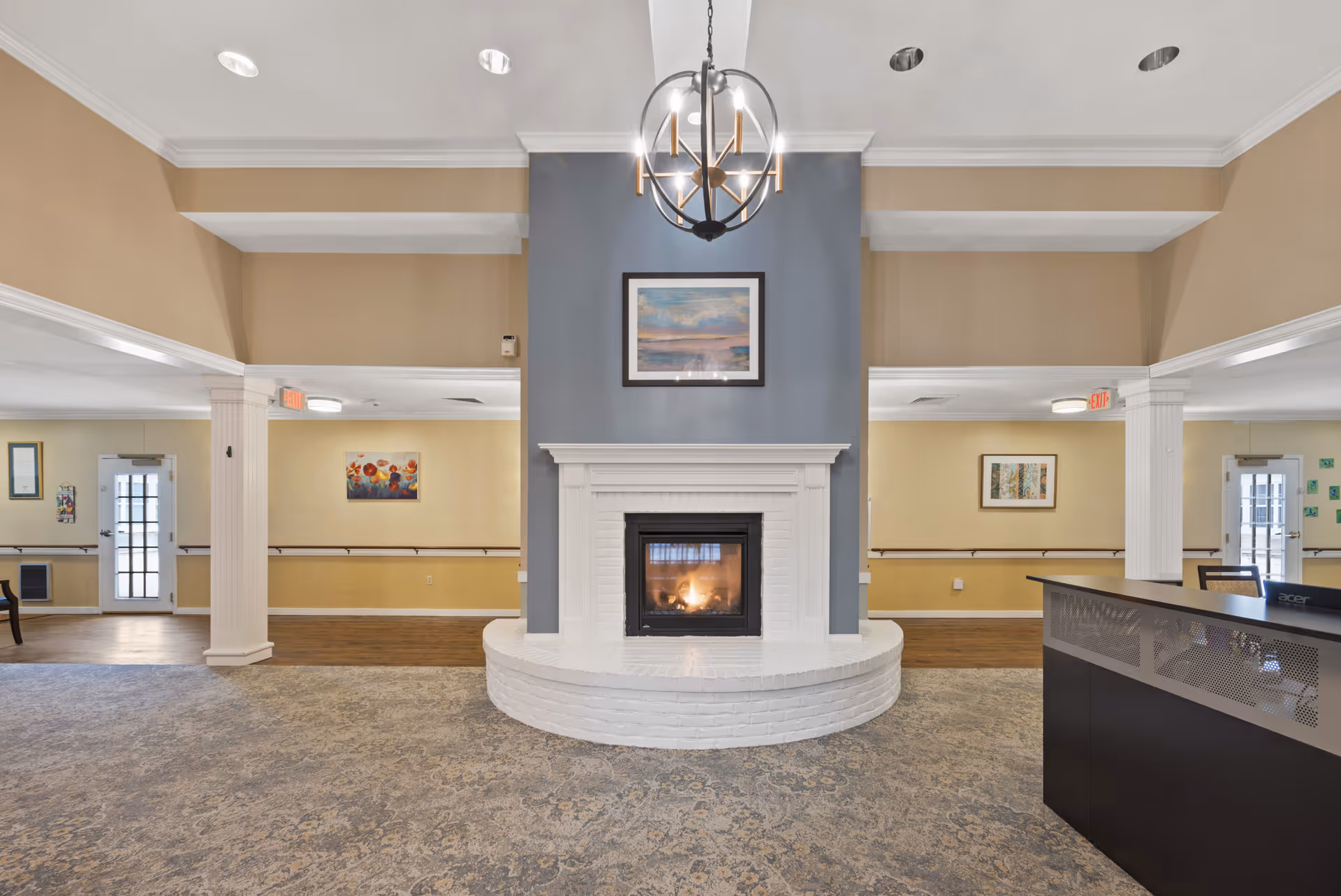 Spacious senior living common area with a central lit fireplace, chandelier, columns and a reception desk.