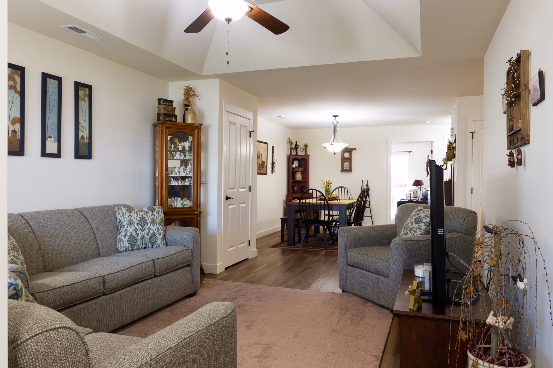 Open living room with gray sofas and an armchair, a ceiling fan, decorative cabinets, and a dining table visible in the background.