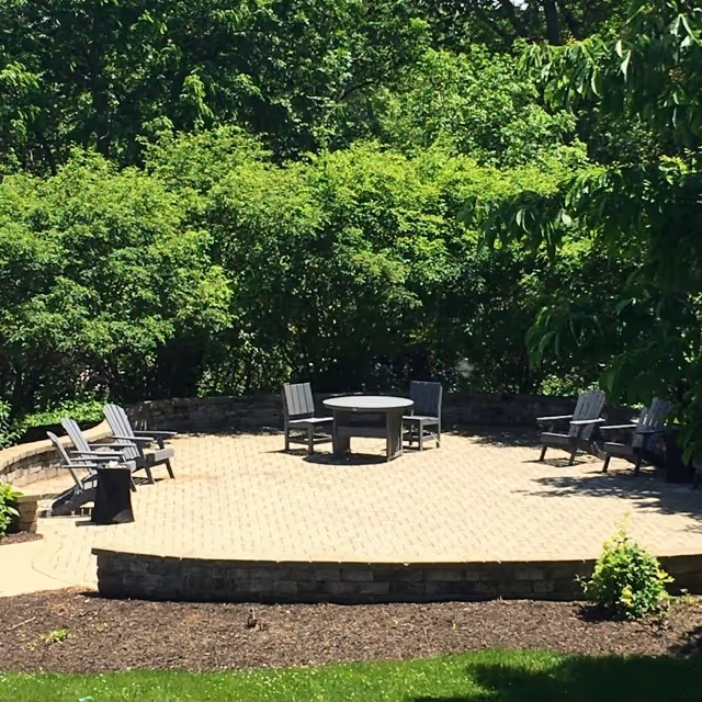 Outdoor patio area with a circular stone fire pit surrounded by six wooden chairs, set on a paved surface with a low stone wall and lush green trees and bushes in the background.