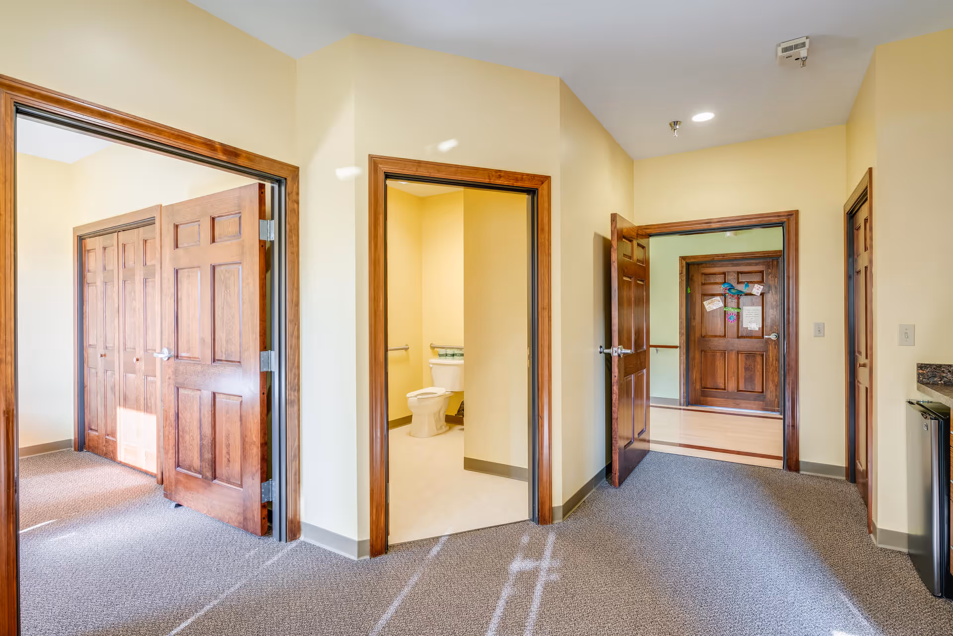 Interior view of a senior living facility hallway with beige walls and carpeted floors. There are three open wooden doors leading to different rooms, including a bathroom with a visible toilet and a room with wooden closets. The hallway is well-lit with natural light and ceiling lights.