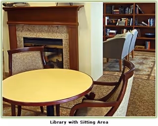Interior view of a senior living community library with a round table and upholstered chairs in the foreground, a fireplace with a wooden mantle on the left, and bookshelves filled with books in the background. The room has carpeted flooring and a cozy, inviting atmosphere.