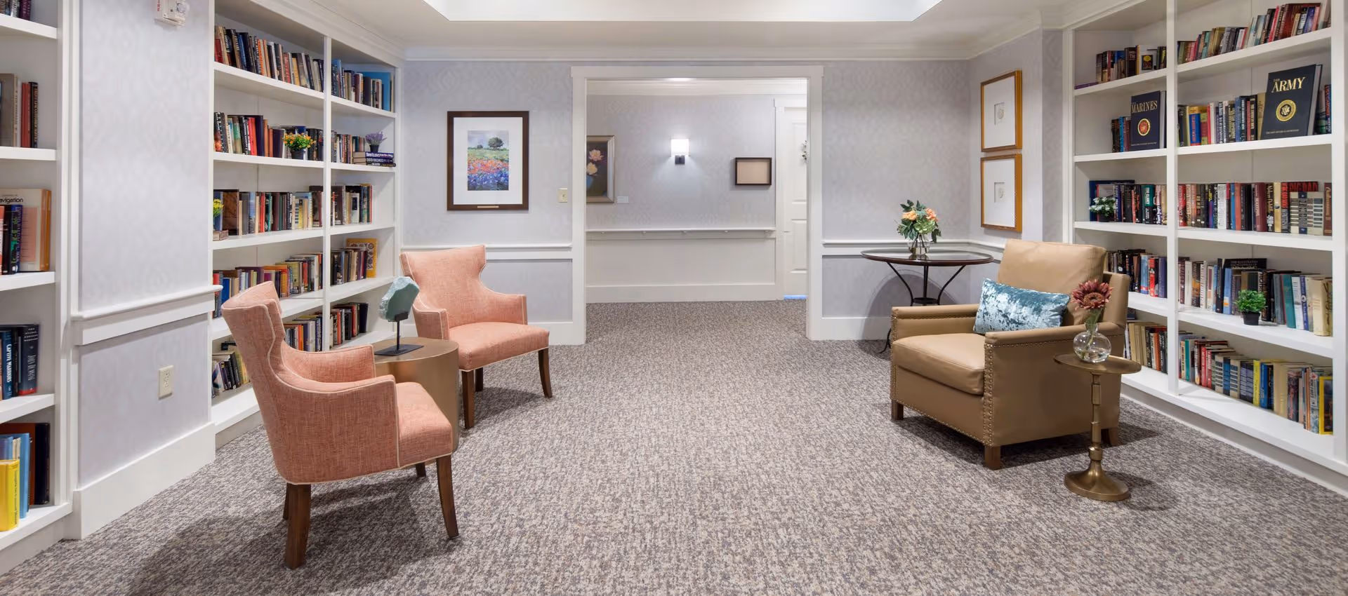 Carpeted library-style sitting area with bookshelves lining the walls, two pink armchairs, a leather armchair and small side tables.