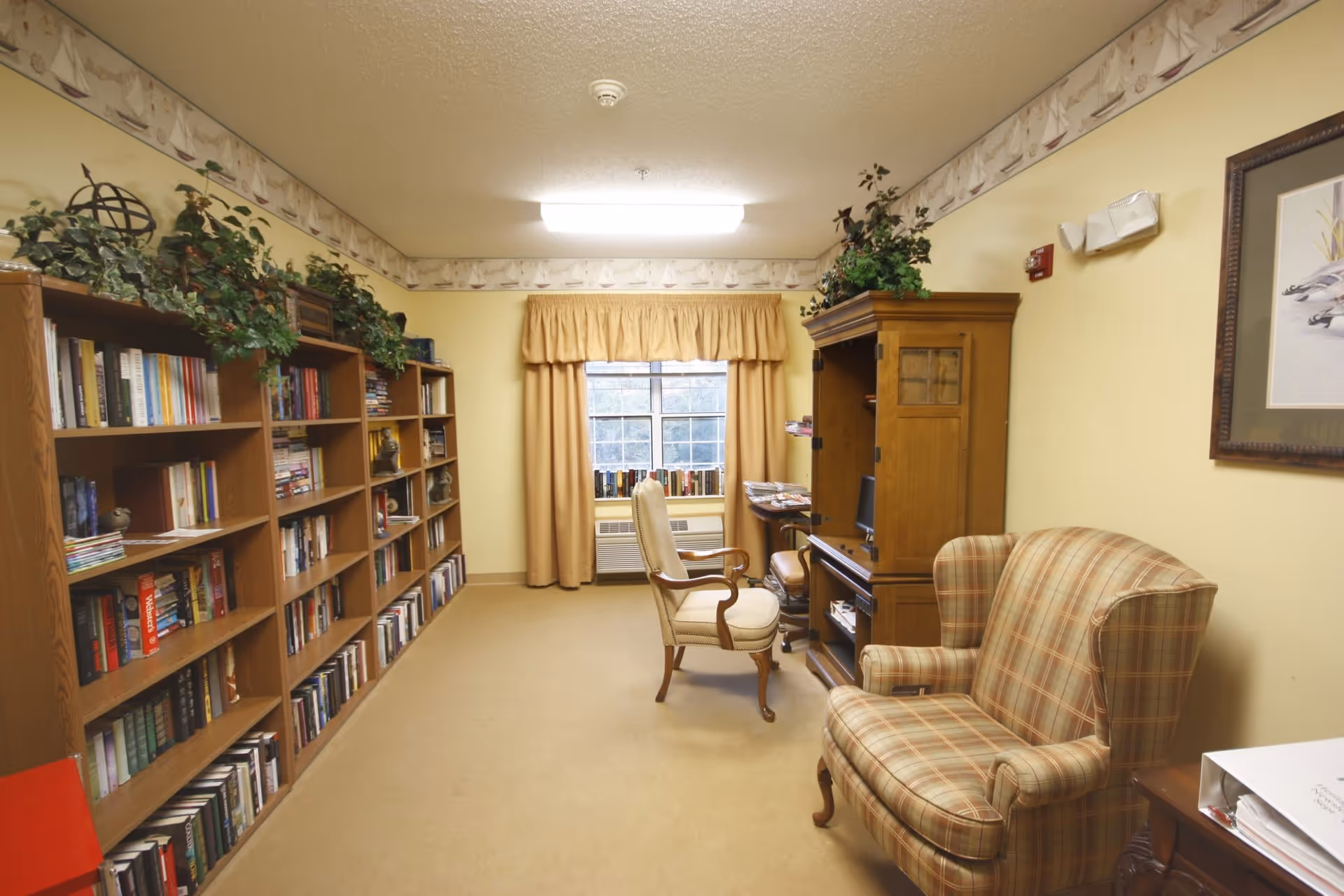 A cozy reading room with bookshelves filled with books along the left wall, a window with beige curtains at the far end, a wooden desk with a computer and chair, and a plaid upholstered armchair on the right side. The walls are painted yellow with a decorative border near the ceiling, and there are green plants on top of the bookshelves and desk.