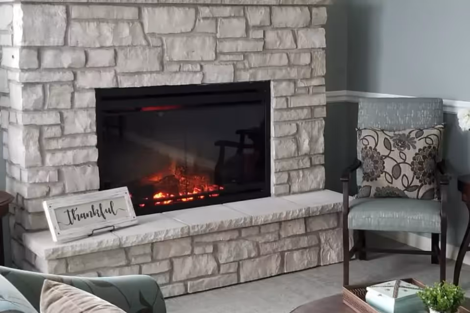 A cozy living room corner featuring a stone fireplace with a glowing fire, a decorative sign that says 'thankful' placed on the hearth, a cushioned armchair with a floral patterned pillow, and part of a coffee table with a small plant and books.