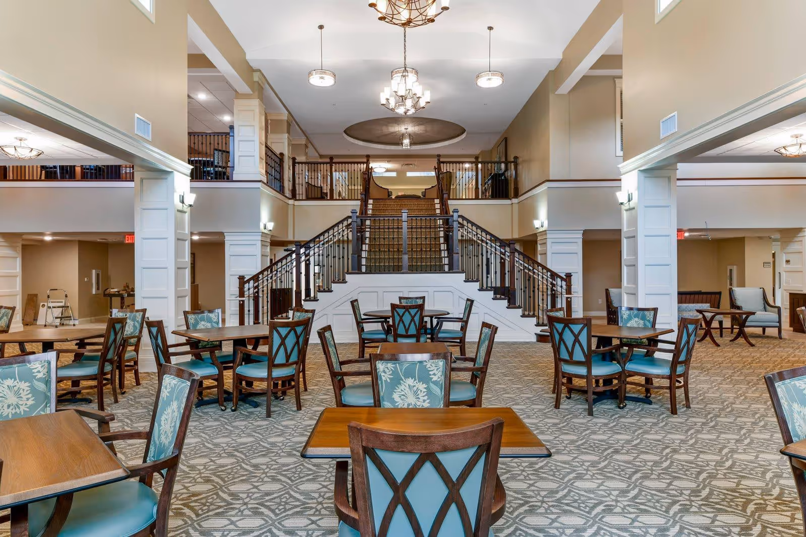 Spacious interior of a senior living facility with multiple wooden tables and chairs featuring teal cushions and floral patterns. A grand staircase with dark wooden railings leads to an upper level with a balcony. The room has high ceilings with elegant chandeliers and beige walls accented by white columns and trim.
