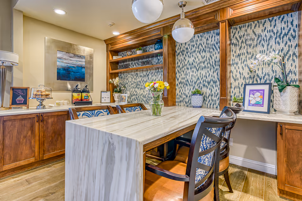 A cozy dining area with a marble table surrounded by wooden chairs with patterned cushions. The background features wooden cabinetry with decorative blue and white patterned panels, a vase with flowers on the table, and various decorative items including framed pictures and plants. There is also a counter with a lamp, a display of pastries, and beverage dispensers.