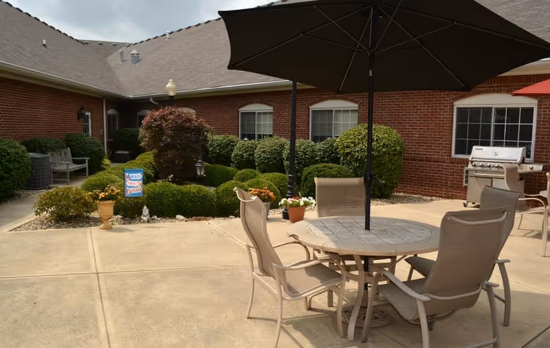 Outdoor patio area with a round table and four chairs under a large black umbrella. The patio is surrounded by well-maintained bushes and plants, with a brick building in the background. There is a barbecue grill on the right side and a bench on the left side near the bushes.