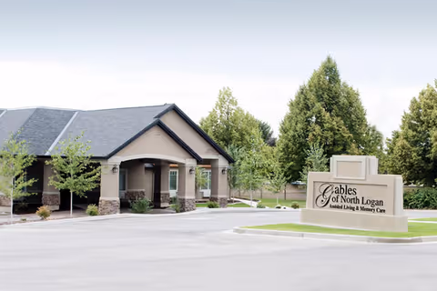 Exterior view of The Gables Assisted Living of North Logan building with a driveway and landscaped trees. A large sign in front reads 'Gables of North Logan Assisted Living & Memory Care'.