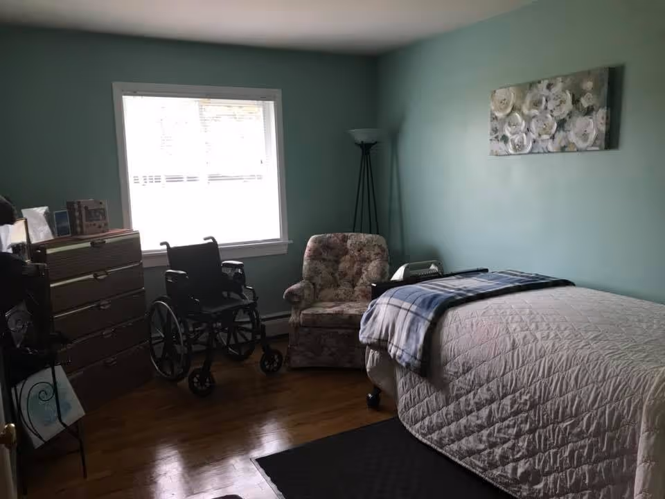 A bedroom with a single bed covered with a beige quilt and a blue plaid blanket folded at the foot. There is a floral armchair next to the bed, a black wheelchair near a window with blinds, a tall floor lamp, and a wooden dresser with several items on top. The walls are painted light green and there is a floral painting hanging above the bed.