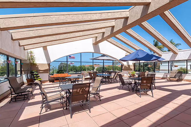 A spacious outdoor patio area with multiple tables and chairs arranged for seating. The patio is covered by a pergola-style roof with large beams and has glass walls on the sides. There are umbrellas providing shade over some tables, potted plants, and an American flag visible outside. The sky is clear and blue.