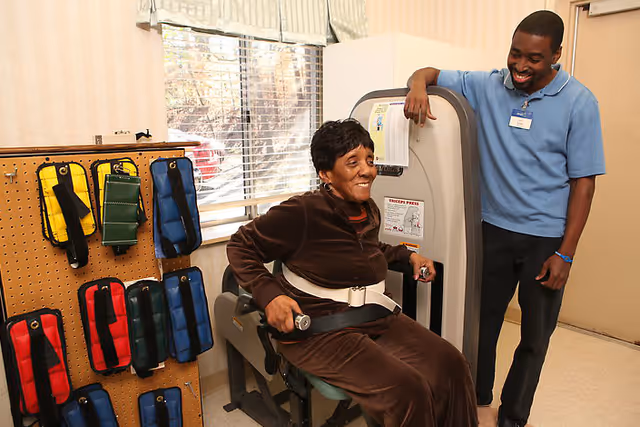 An elderly woman using a triceps press exercise machine in a room with a window and a pegboard holding colorful exercise weights. A male staff member in a blue shirt stands beside her, smiling and providing assistance.
