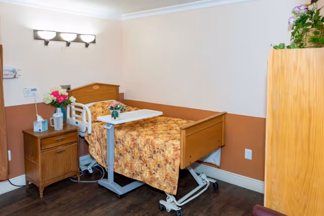A small patient room with a hospital bed covered in a floral patterned blanket. Next to the bed is a wooden nightstand with a tissue box, a water pitcher, and a vase of flowers. A small overbed table with a flower vase is positioned over the bed. The walls are painted half brown and half beige, and there is a wooden cabinet with plants on top in the corner.