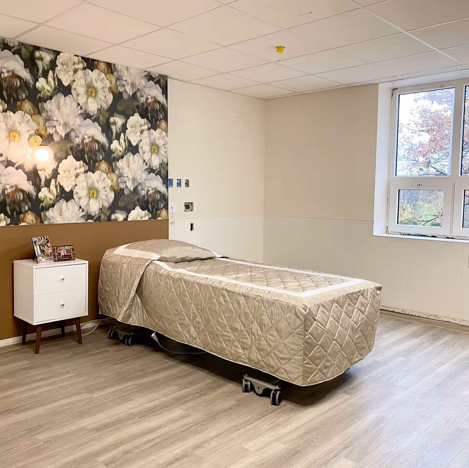 A simple room with a single bed covered in a beige quilted blanket on wheels, a small white nightstand with two drawers holding framed photos, a floral wallpaper accent wall with large white flowers, a wall-mounted light fixture, and a large window letting in natural light.