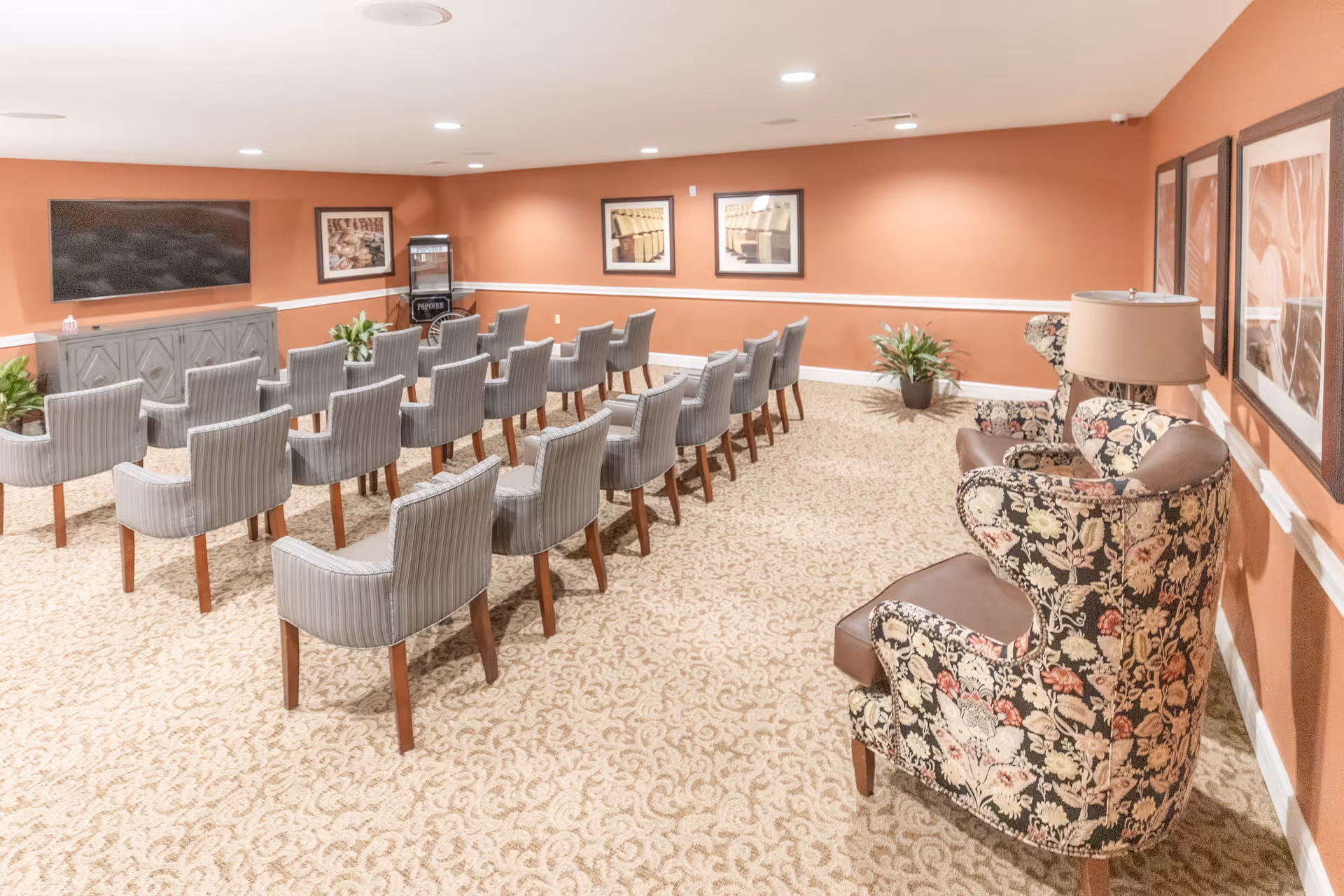 A senior living facility common room with rows of gray upholstered chairs facing a large flat-screen TV mounted on a peach-colored wall. The room has patterned carpet, floral armchairs with a lamp on a side table, framed pictures on the walls, and potted plants.