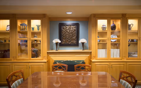 A well-lit room with wooden built-in cabinets displaying decorative items such as vases, books, and sculptures. In the center is a wooden table with chairs around it, and a fireplace with a decorative wall art piece above it flanked by two floral arrangements.