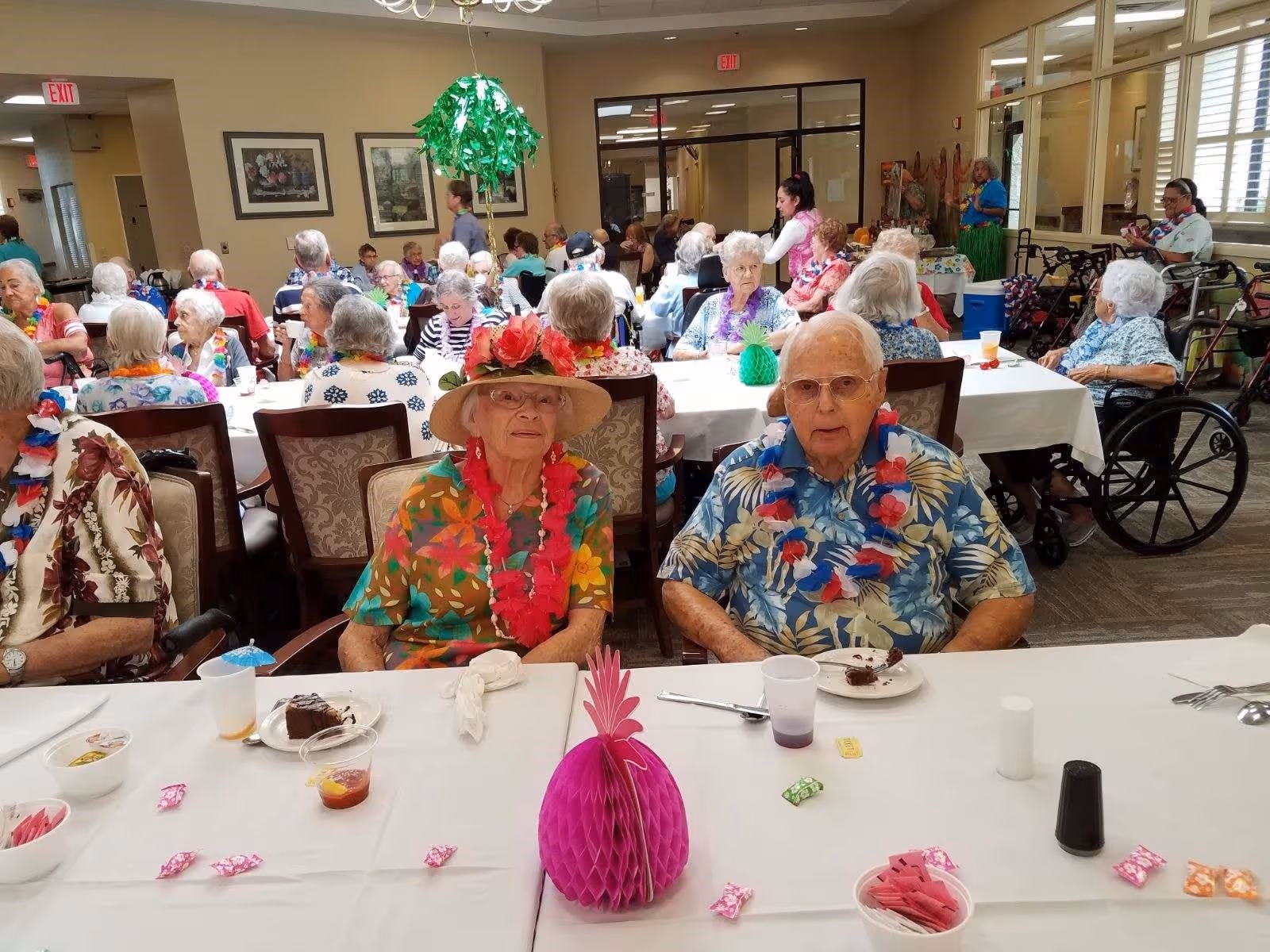 A group of elderly people gathered in a dining room decorated with tropical-themed accessories. Two elderly individuals sit at the front table wearing colorful Hawaiian shirts and leis, with plates of dessert in front of them. Other seniors are seated at tables in the background, some in wheelchairs, enjoying the social event. The room has beige walls, framed pictures, and large windows.