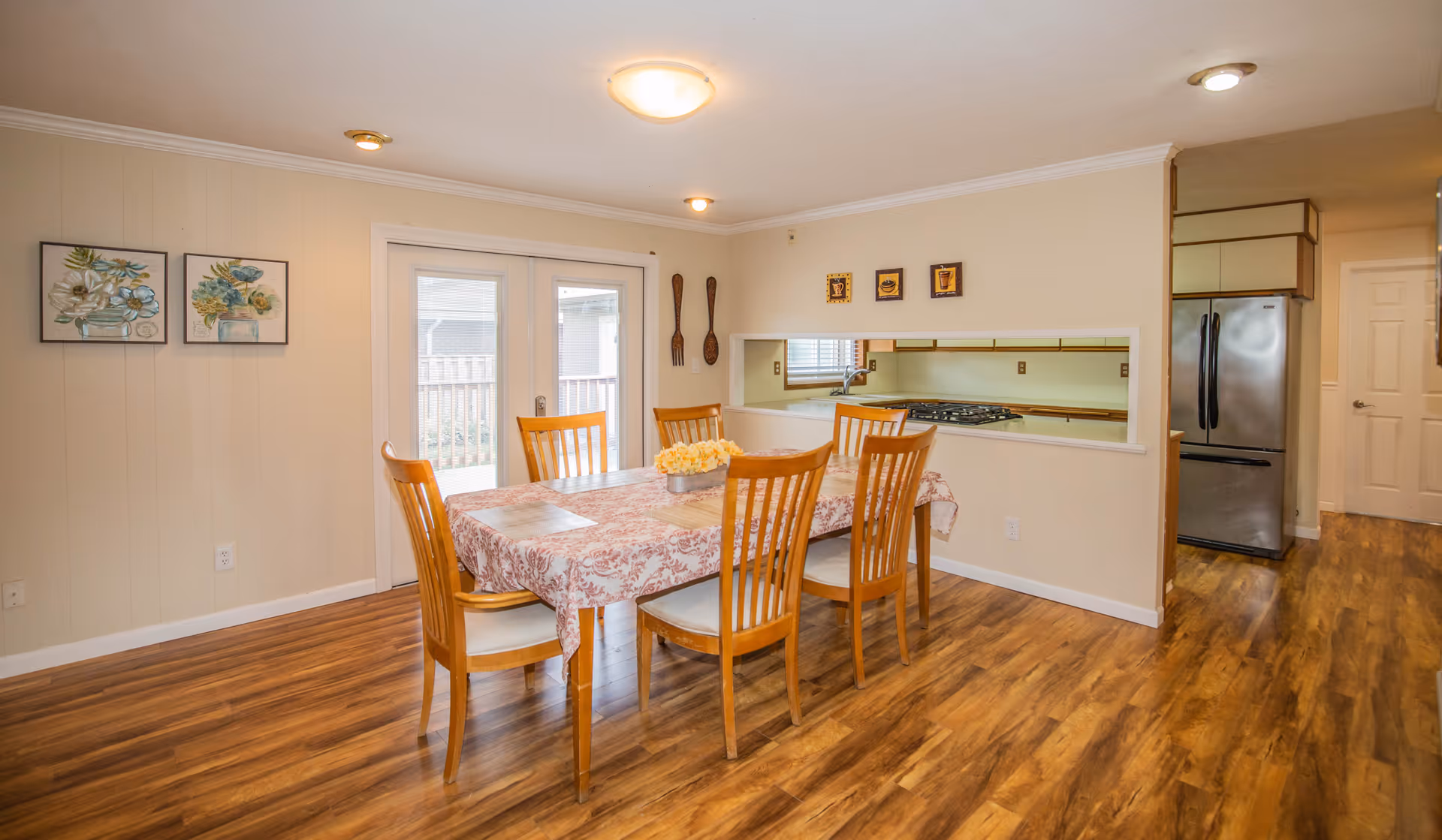 A dining room with a wooden table covered by a floral tablecloth and six wooden chairs. The room has wood flooring, beige walls, and two framed floral paintings on the left wall. There are double glass doors leading outside, and a pass-through window to the kitchen where a stainless steel refrigerator and stove are visible.
