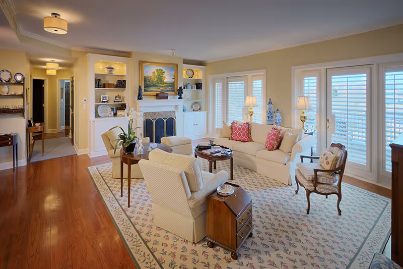 A bright and cozy living room with hardwood floors, a large patterned area rug, a white sofa with red and beige pillows, two upholstered armchairs, a wooden side table, and a fireplace with built-in shelves on either side. The room has large windows with white plantation shutters and two table lamps providing warm lighting.