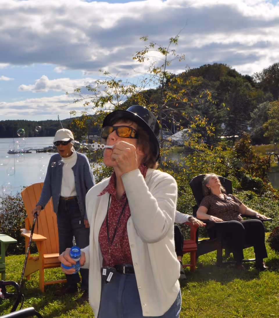 An elderly woman wearing a black hat and sunglasses blows bubbles outdoors near a lake, with another elderly woman standing nearby using a cane and a third woman relaxing in a chair under a partly cloudy sky.