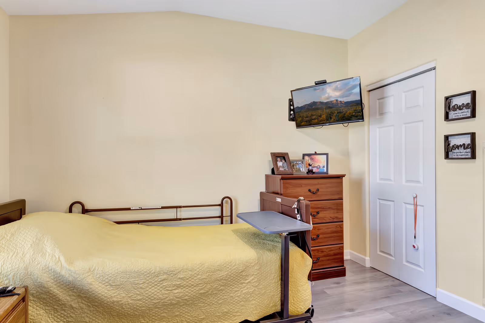 A senior living bedroom with a bed covered in a yellow quilt, a wooden dresser with framed photos on top, a wall-mounted TV displaying a scenic landscape, and two decorative wall plaques with the words 'love' and 'home'. There is a white closet door with a hanging emergency call button.