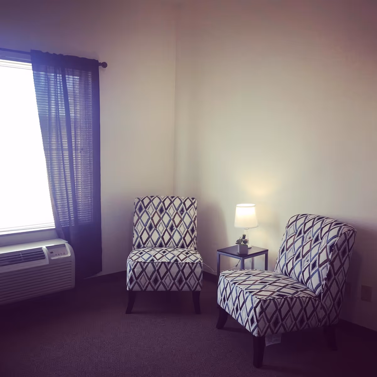 A corner of a room with two patterned armchairs facing slightly towards each other, a small side table with a lit lamp and a small plant, a window with dark curtains, and an air conditioning unit below the window.