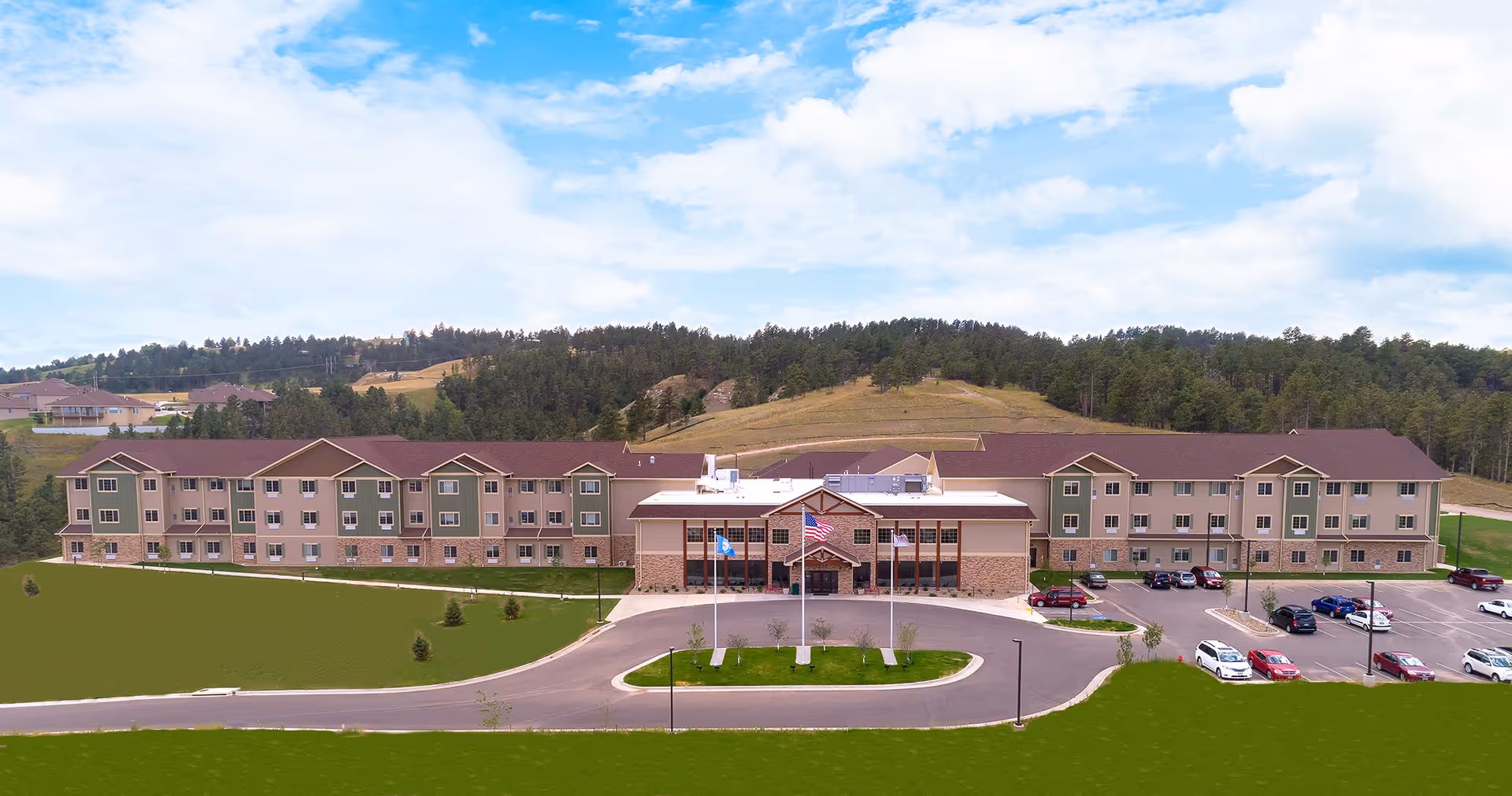 Wide exterior view of The Village at Skyline Pines Assisted Living & Memory Care building, a large three-story facility with a brown roof and beige and green walls, surrounded by a parking lot and green lawns, set against a backdrop of forested hills under a partly cloudy sky.
