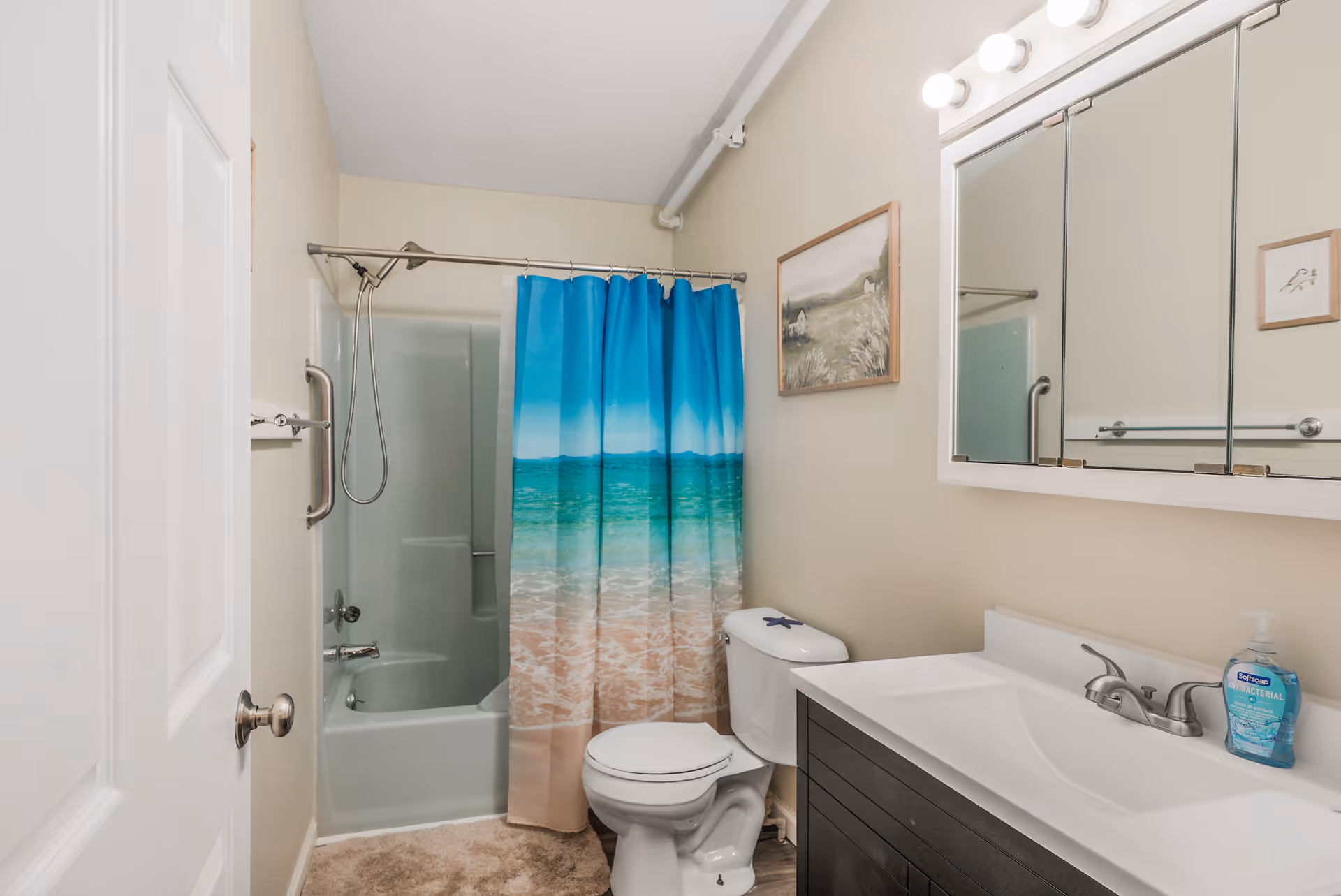 A clean bathroom featuring a bathtub/shower with a blue beach-themed curtain, a toilet, and a vanity with sink and mirrored medicine cabinet.