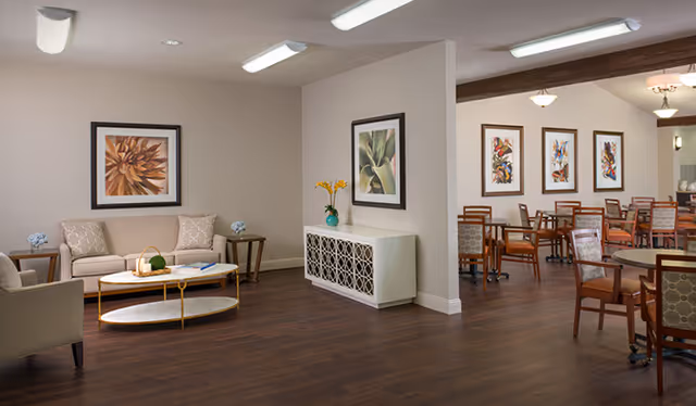 Interior view of a senior living facility showing a seating area with a beige sofa, armchair, coffee table, and side tables with decorative items. Adjacent to this is a dining area with multiple wooden tables and chairs, and framed artwork on the walls. The flooring is dark wood, and the lighting is provided by ceiling fixtures.