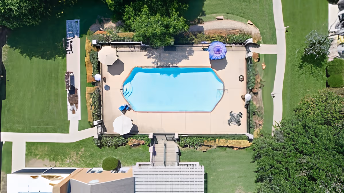 Aerial view of an outdoor swimming pool area surrounded by a concrete deck with several umbrellas and lounge chairs. The pool area is bordered by green lawns, trees, and pathways leading to and from the pool. Part of a building is visible at the bottom of the image.