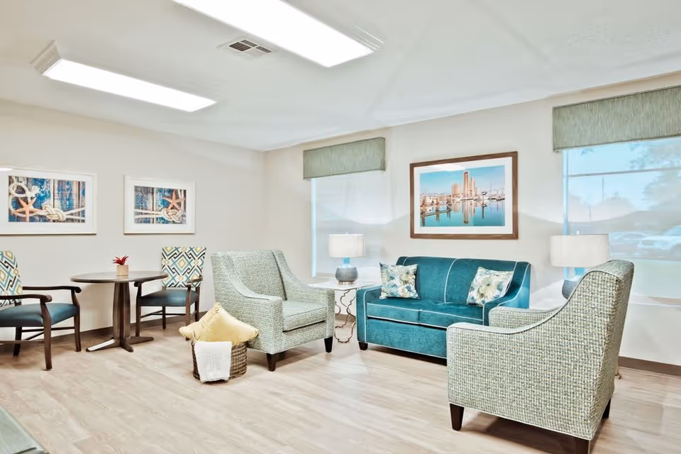 A bright and cozy living room area in a senior living facility featuring a teal sofa with floral pillows, two patterned armchairs, a small round table with two chairs, two side tables with lamps, and framed artwork on the walls. The room has large windows with green valances and light wood flooring.