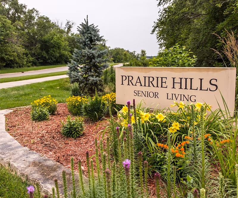 A landscaped garden area with various colorful flowers and plants surrounding a stone sign that reads 'PRAIRIE HILLS SENIOR LIVING'. A road and trees are visible in the background under an overcast sky.