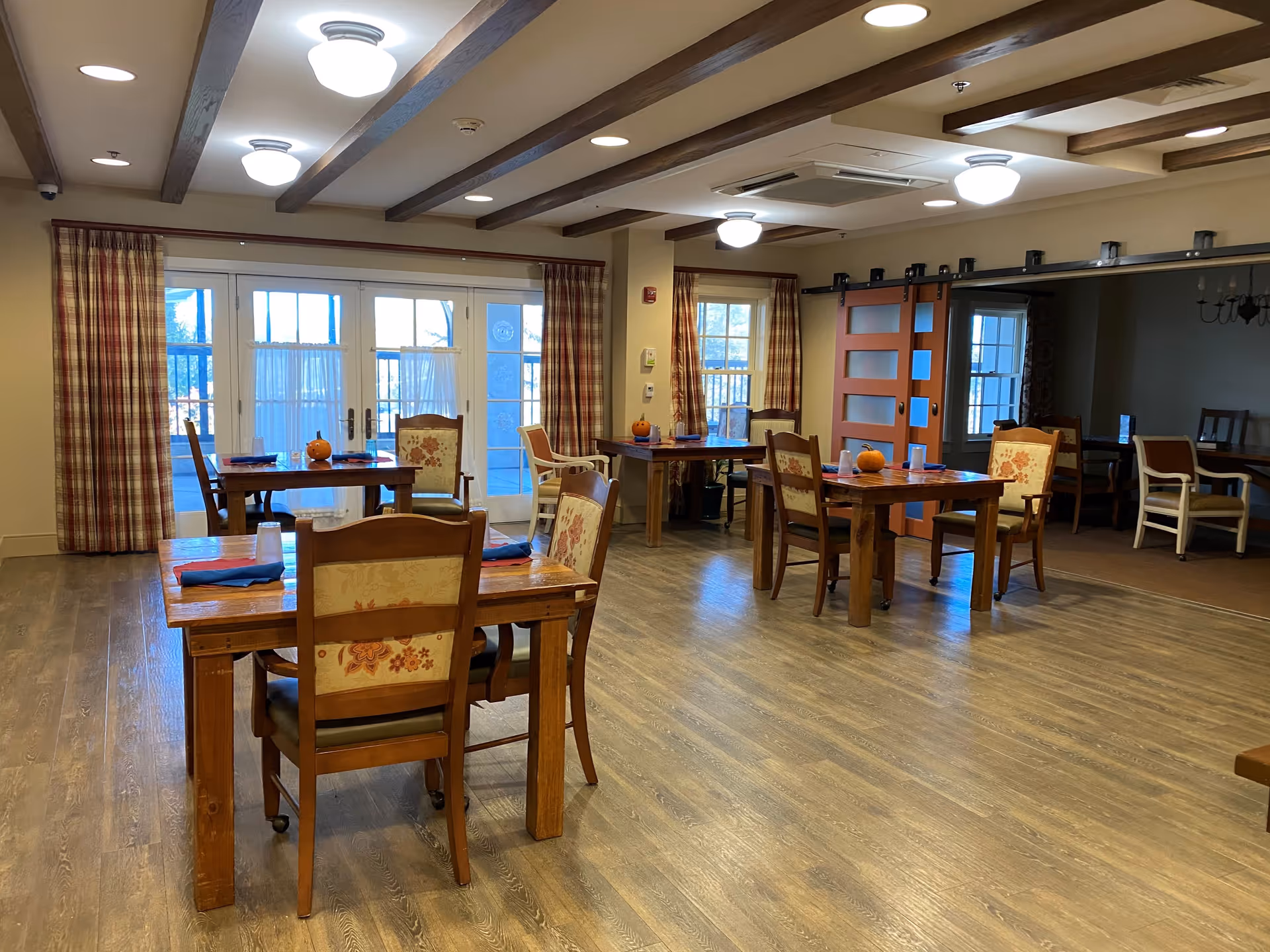 Dining room with wooden tables and upholstered chairs, plaid curtains, and glass doors letting in daylight.