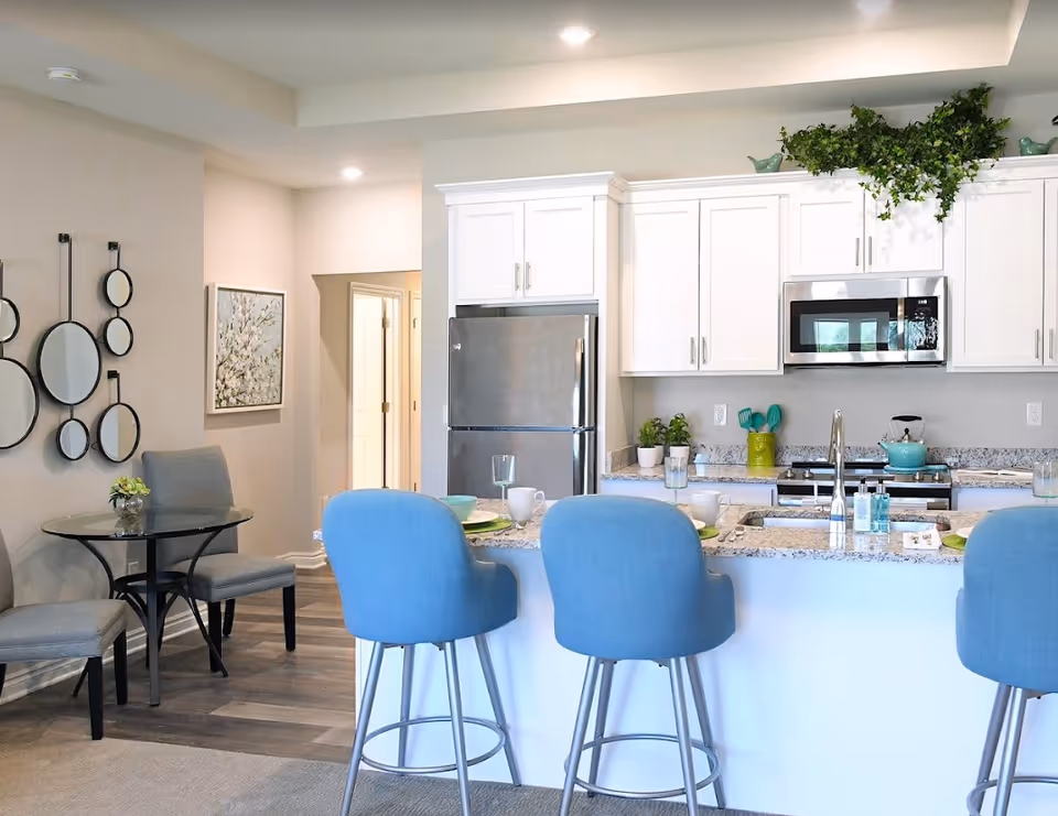 A modern kitchen and dining area with white cabinets, a stainless steel refrigerator and microwave, and a granite countertop island with three blue bar stools. The kitchen counter has place settings with plates, bowls, glasses, and mugs. To the left, there is a small round glass table with two gray upholstered chairs, a wall decorated with multiple round mirrors, and a framed floral painting. The floor is a combination of wood and carpet.