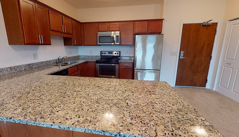 A kitchen with granite countertops, wooden cabinets, a stainless steel refrigerator, stove, and microwave. There is a double sink on the left side and a wooden door in the background.