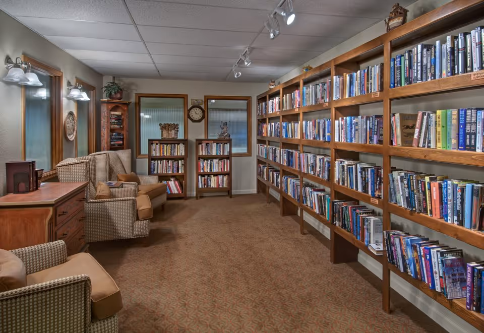 Cozy library room with wooden bookshelves lining the right wall, armchairs and small tables on the left, and a carpeted floor.