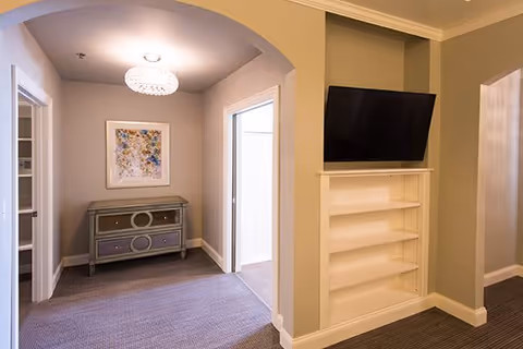 Interior hallway alcove with an arched entry, decorative console and framed artwork, carpeted floor, and a wall-mounted TV above built-in shelves.